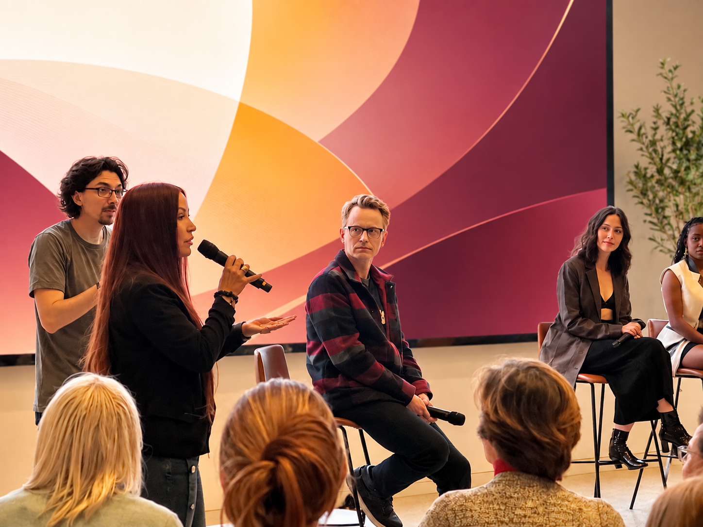 Annie Biggs speaking into a handheld microphone beside seated panelists, audience in the foreground