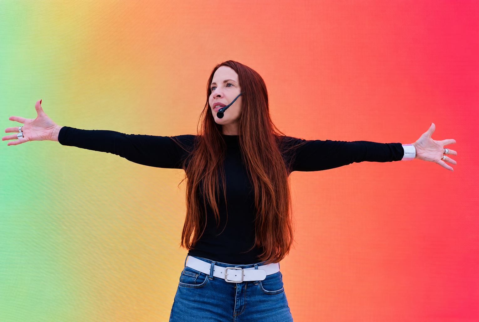 Annie Biggs speaking with a headset microphone, both arms extended against a colorful stage background.